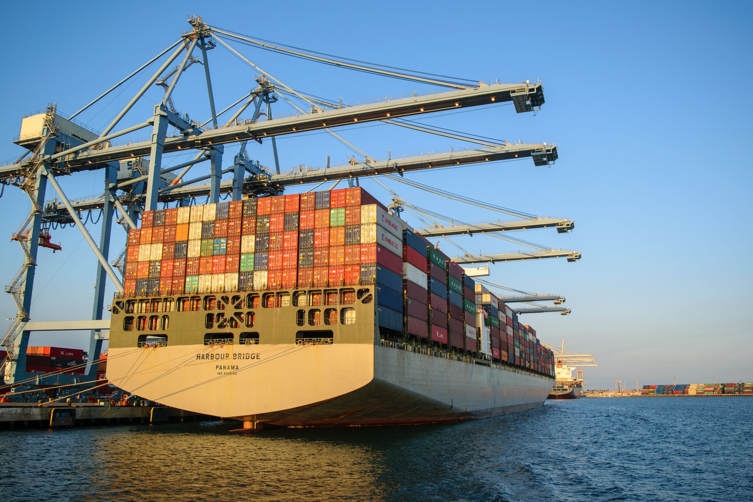 Home A large container ship at an industrial harbor with cranes under a clear blue sky.